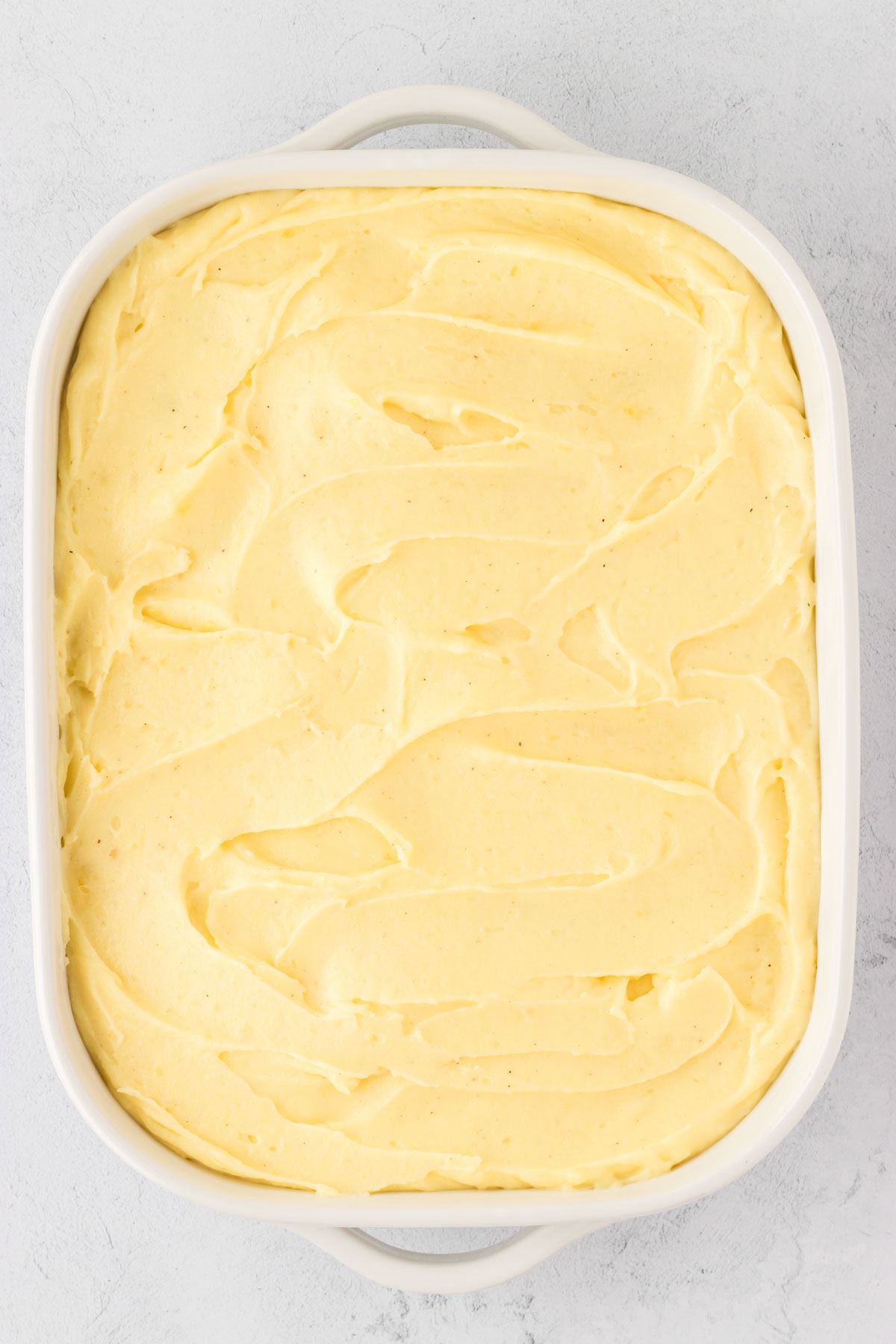 A casserole dish filled with ground beef filling and then adding mashed potatoes smoothed evenly over the top.