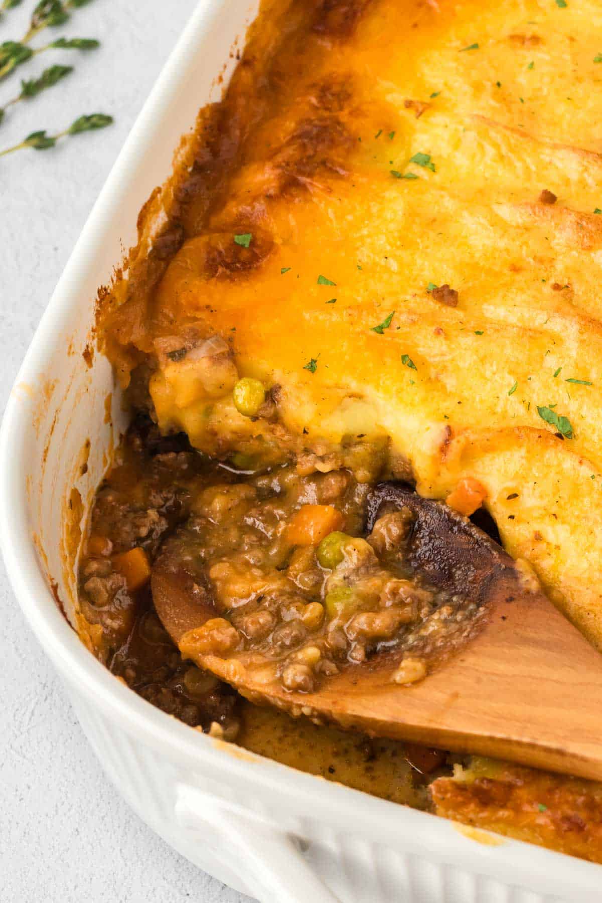 A white casserole dish filled with Shepherd's Pie with a scoop removed with a wooden spoon.