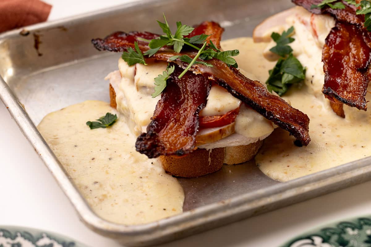 A baking sheet is pictured with a2 slices of Hot Brown sitting smothered in mornay sauce and garnished.