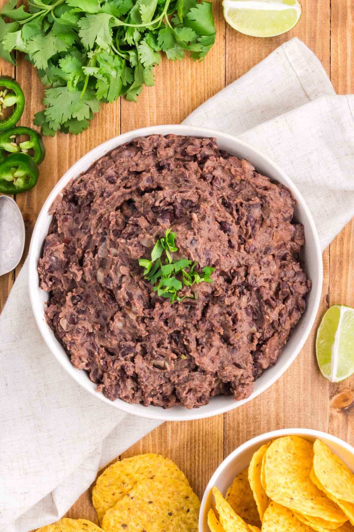 A table is set with a bowl of chips and and other garnishes surrounding a serving bowl of Refried Black Beans.