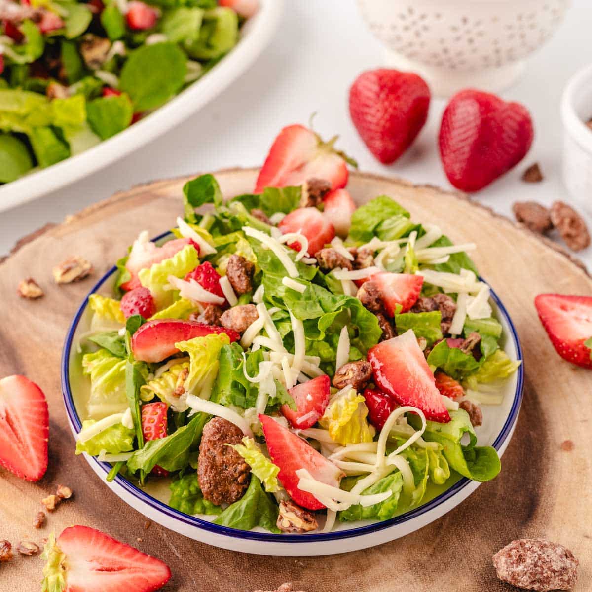 A wooden charger with a white plate with blue rim filled with a strawberry praline pecan salad with mozzarella and honey vinaigrette.