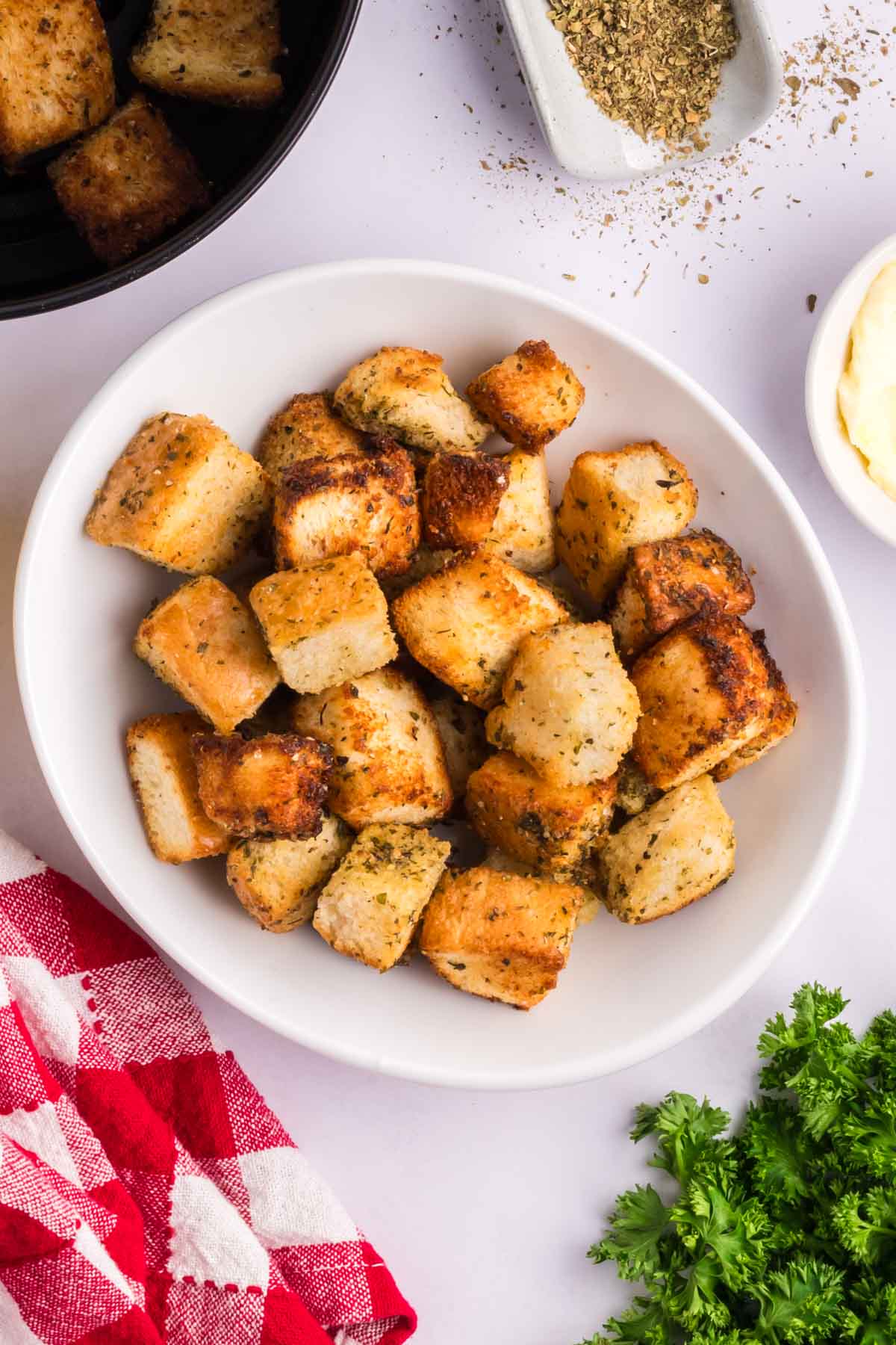 Homemade croutons in a white bowl with a red white checked napkin and bunch of parsley to the side.