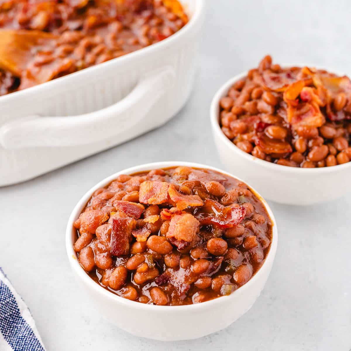 Two bowls of baked beans with casserole serving dish in background.