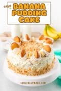 A cake stand sits with a banana pudding cake topped with whipped icing, vanilla wafers and banana slices on top.