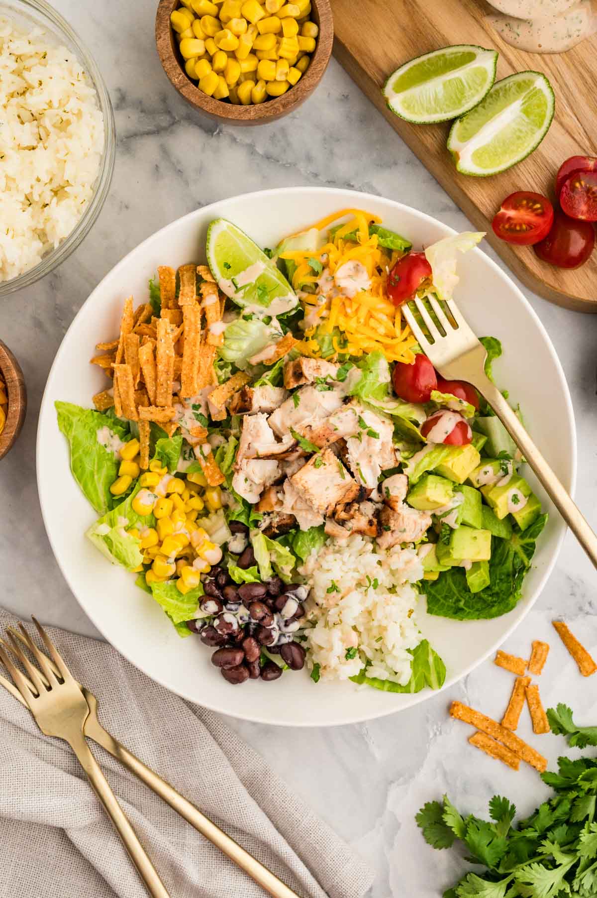 A white serving bowl with a bed of lettuce greens topped with bbq chicken, rice, cheese, corn, black beans, avocado and tomatoes.