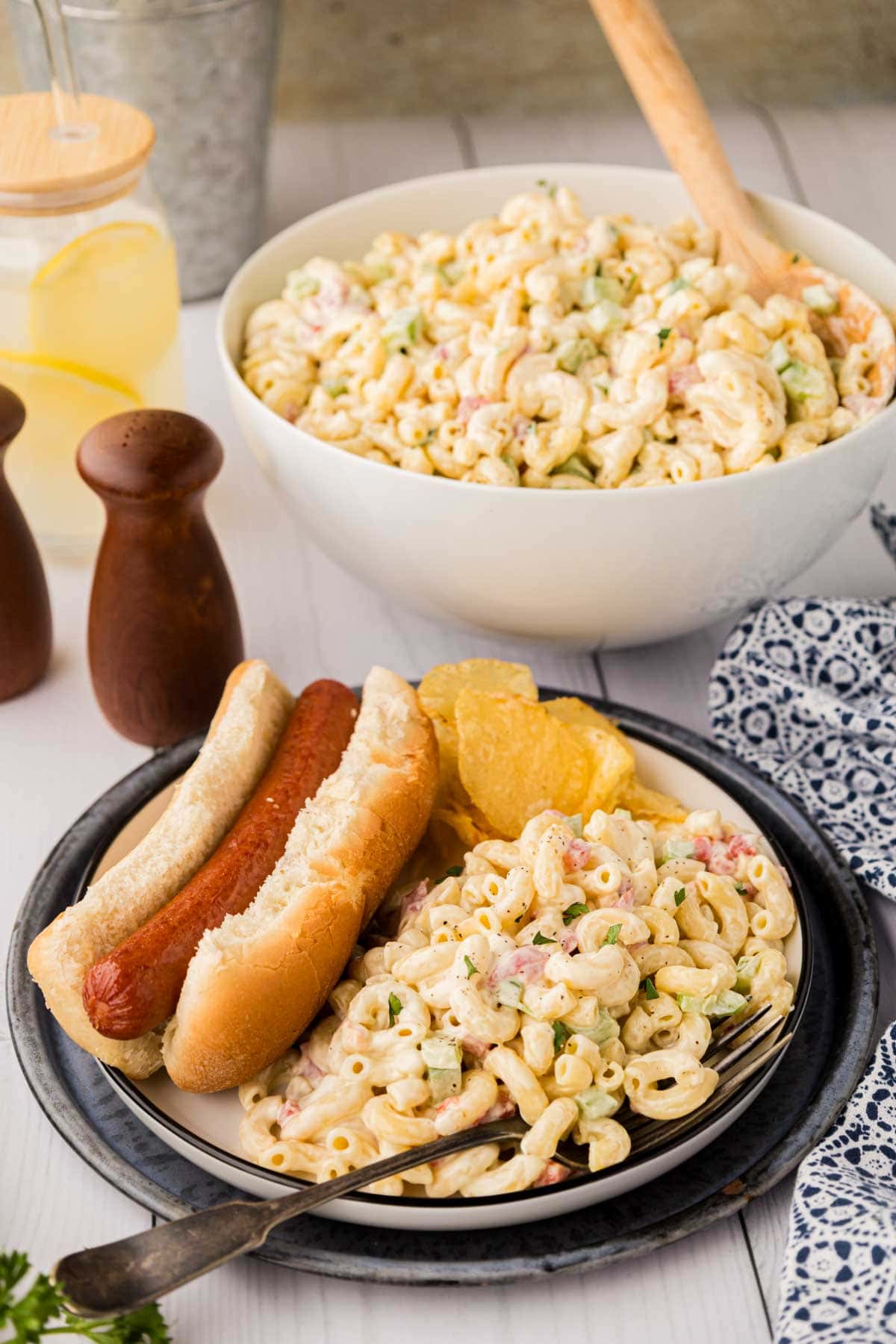 Macaroni salad served with a hot dog on a plate with a bowl of macaroni in the background.