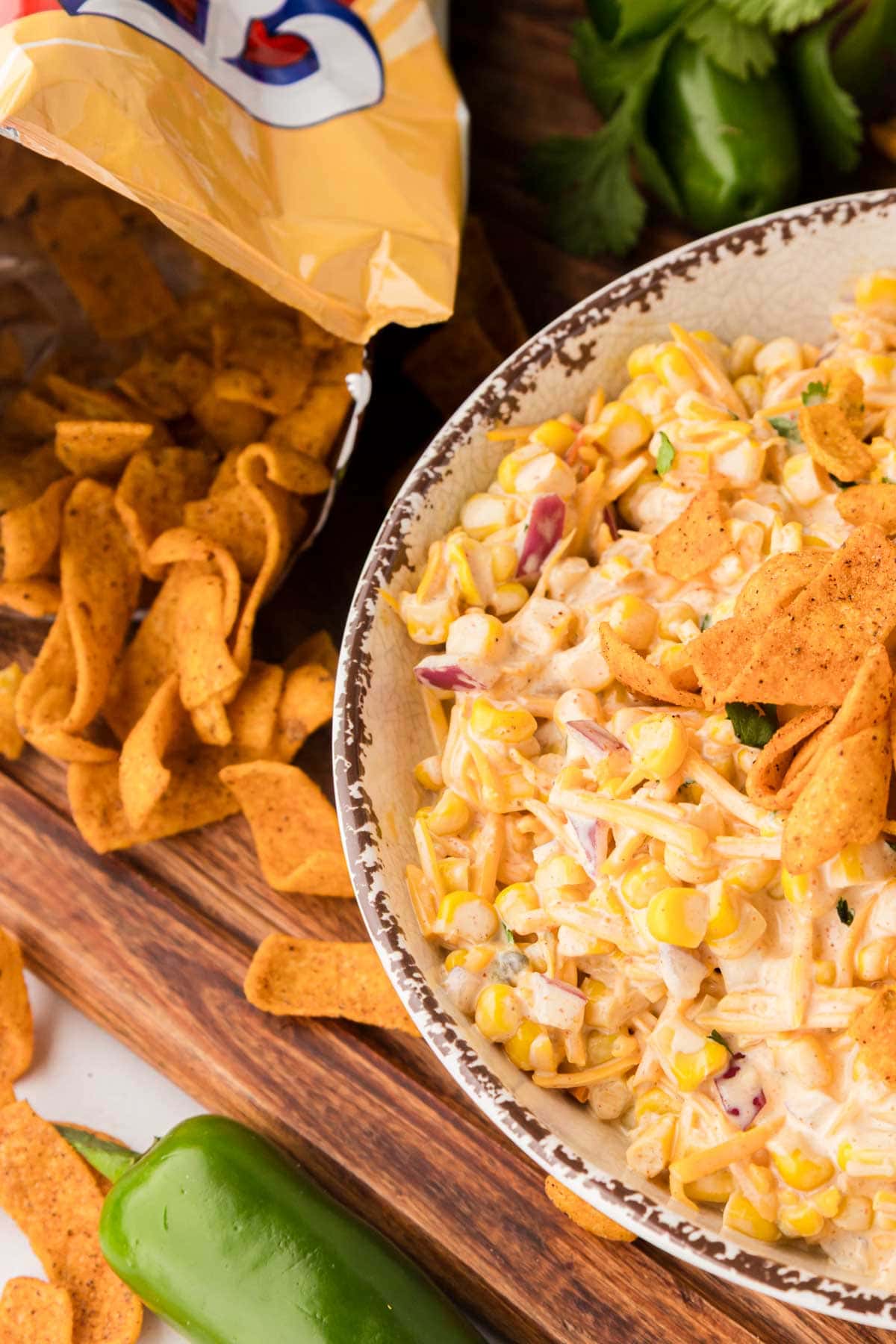 An open bag of fritos with corn salad in a bowl on a wooden board.