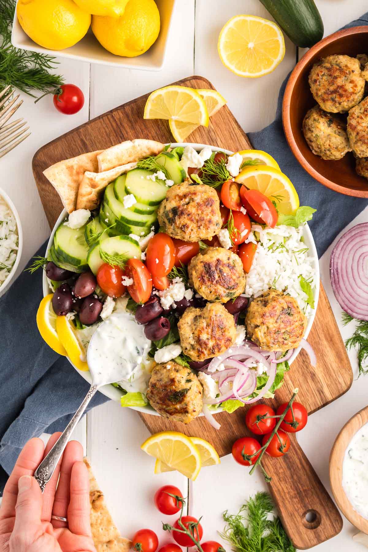 Greek chicken meatballs on a bed of rice and veggies with a hand putting tzatziki sauce on it.