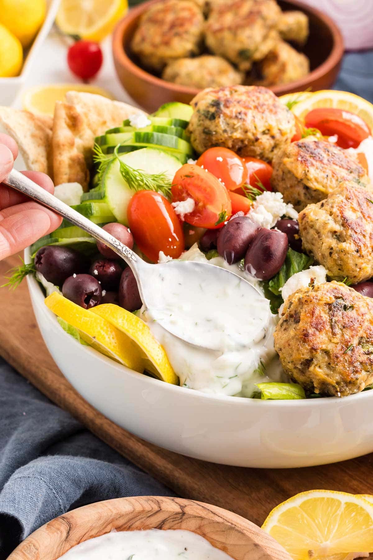 Using a spoon to place tzatziki sauce in a bowl of Greek chicken meatballs and veggies.