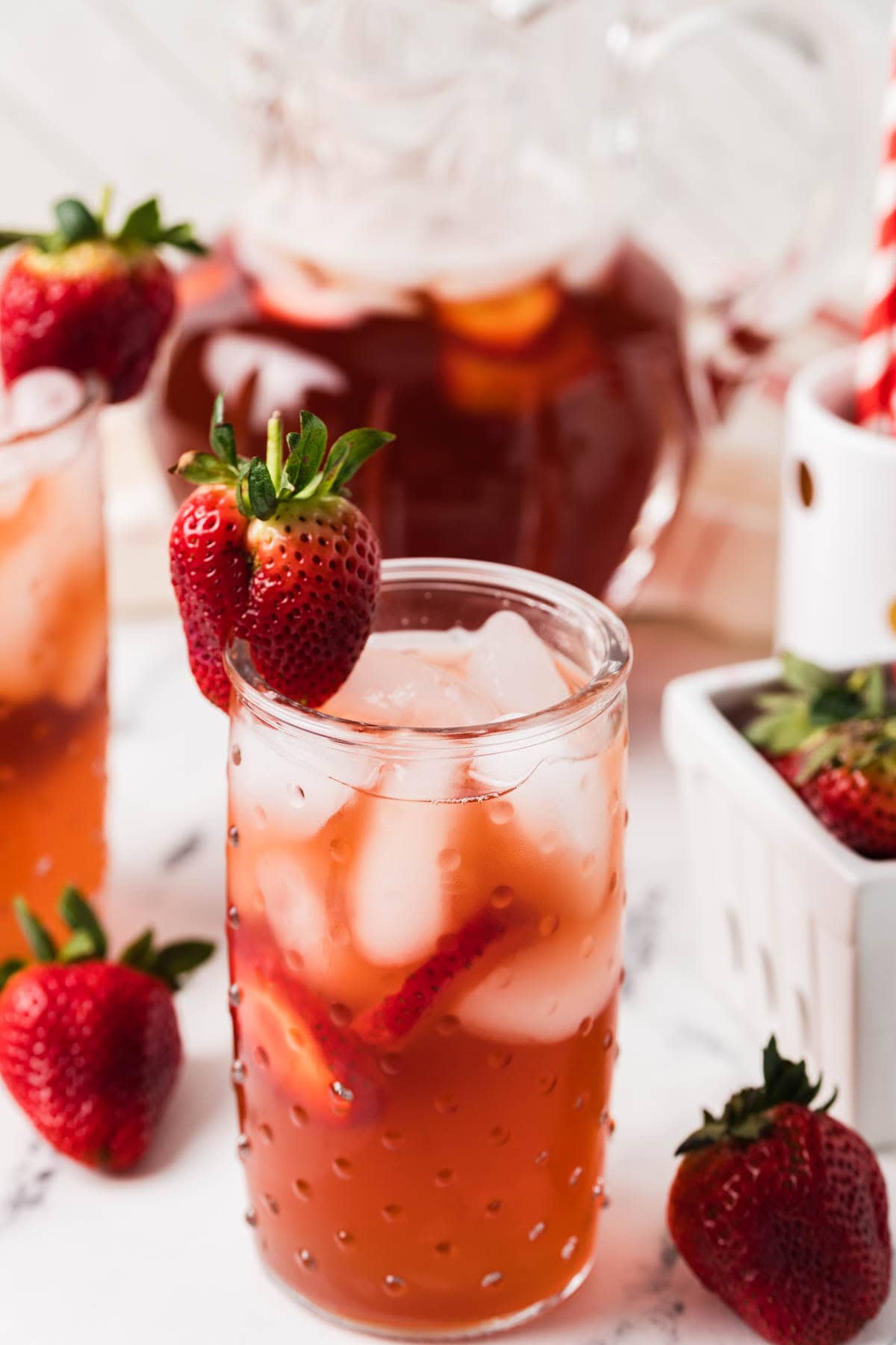 Glass of iced strawberry Southern tea garnished with a fresh strawberry with the pitcher in the background.