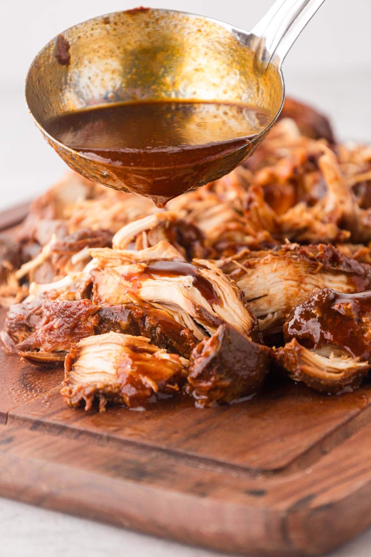 Shredded BBQ chicken on a wooden board, ladling the sauce on top.