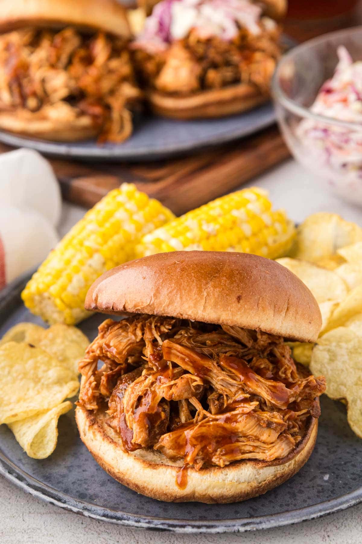 Slow cooker pulled BBQ chicken on a bun served with corn and potato chips.