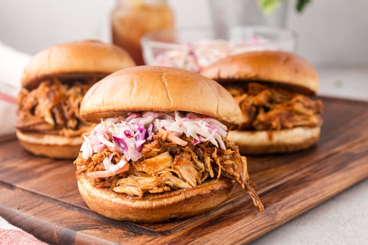 Three pulled BBQ chicken sandwiches on a wooden board.