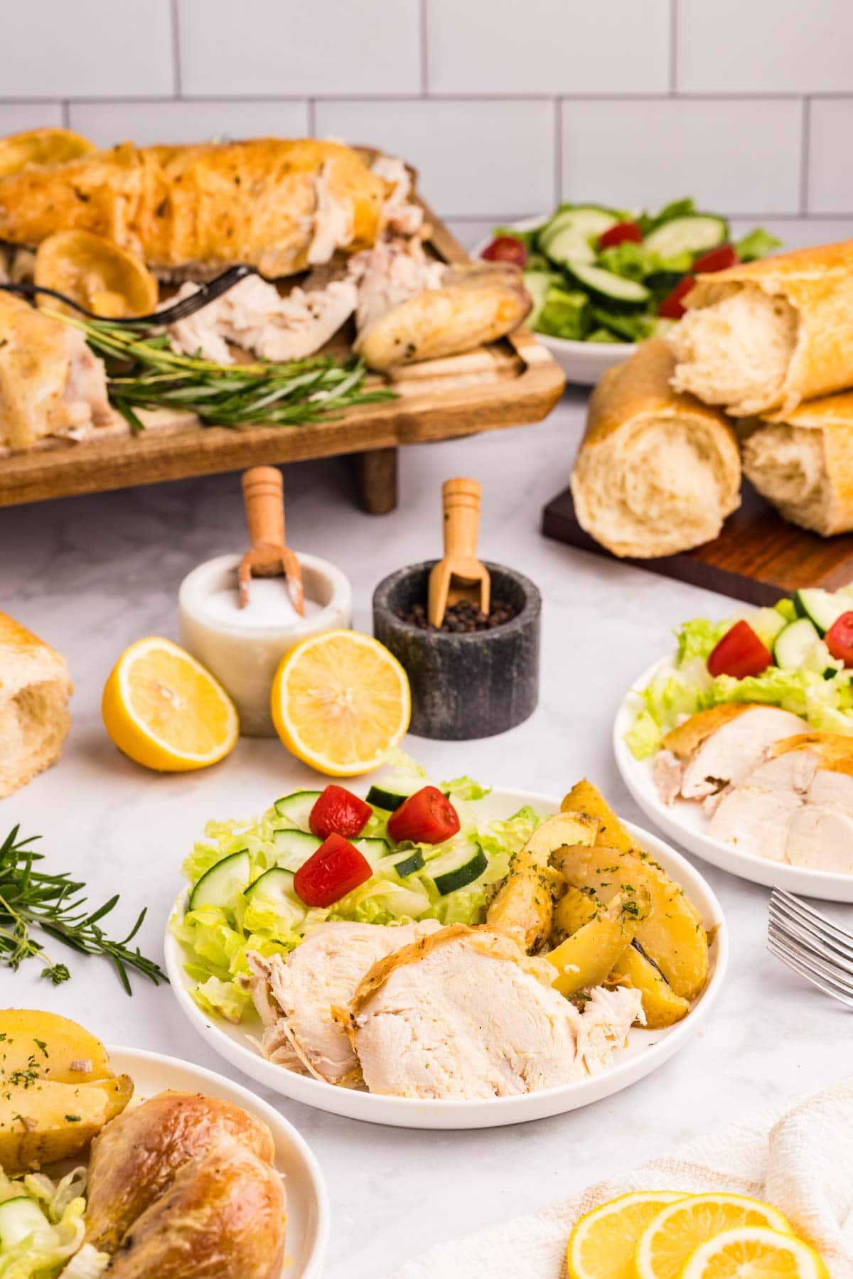 Slices of Greek roast chicken served on a plate with the potatoes, a salad, and more bread and salad around it.
