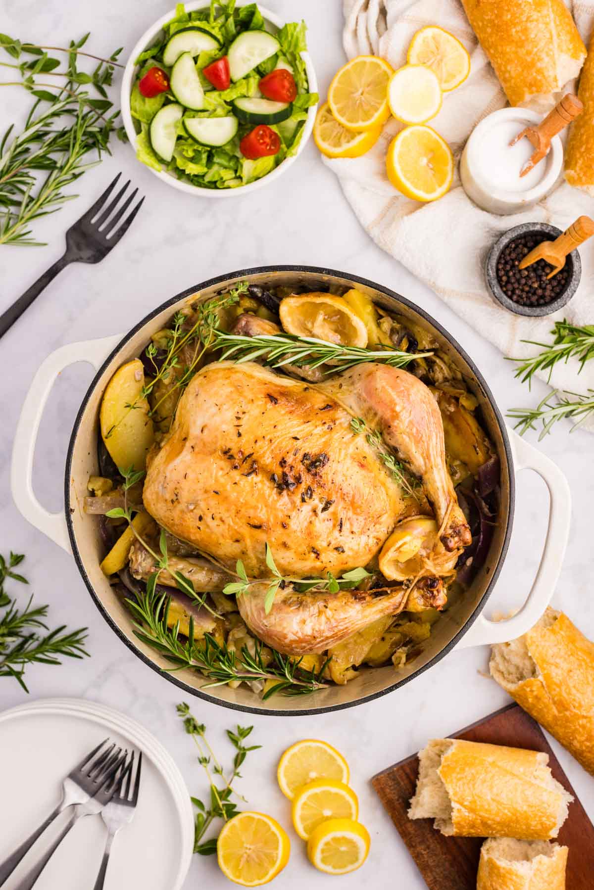 Overhead shot of the Greek roast chicken in a Dutch oven with bread, salad, and herbs around it.