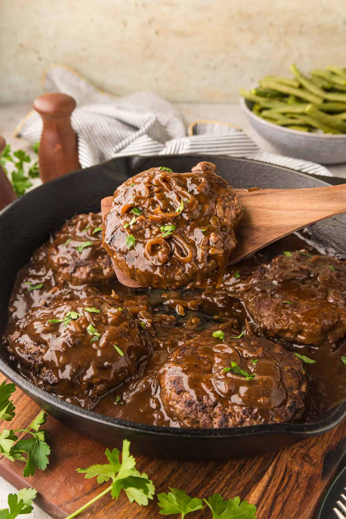 Hamburger steaks with onion gravy in a cast iron skillet with a wooden spoon removing a serving.