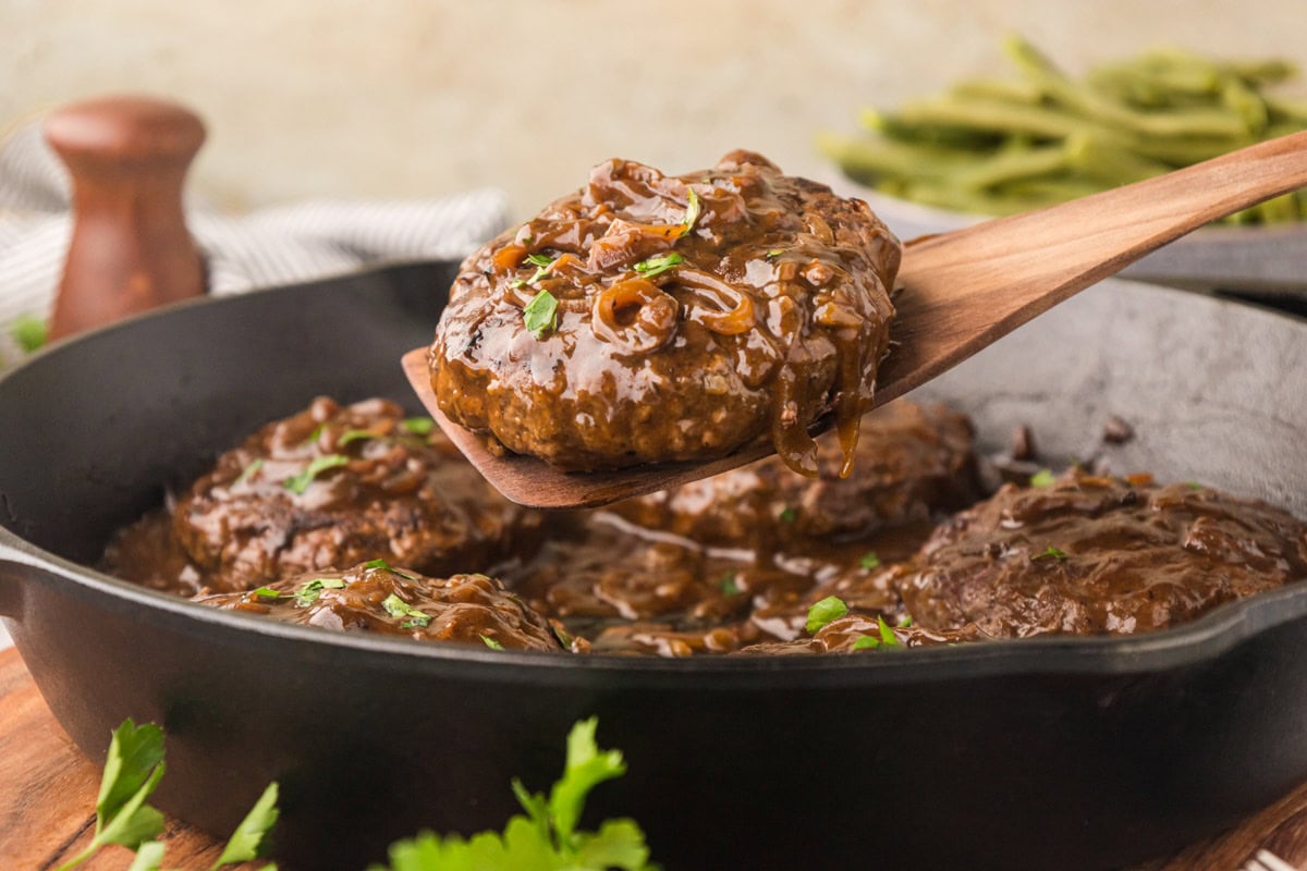 Hamburger steaks with onion gravy in a cast iron skillet with a wooden spoon removing a serving.