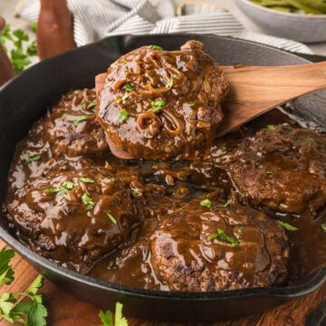 Hamburger steaks with brown gravy in a cast iron skillet with a wooden spoon removing a serving.