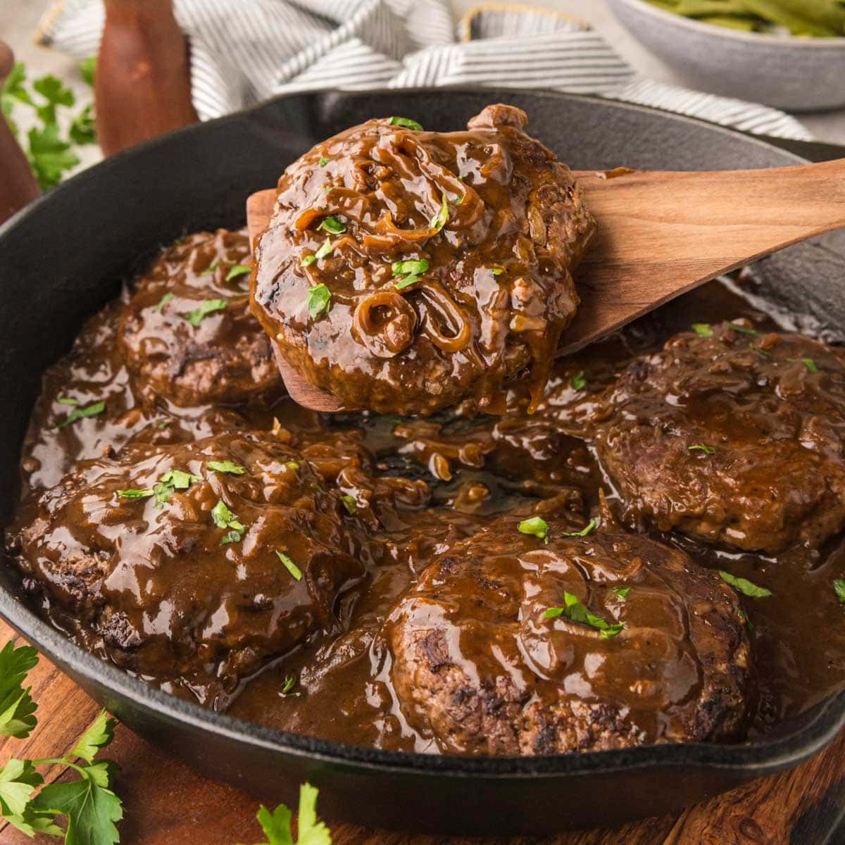 Hamburger steaks with brown gravy in a cast iron skillet with a wooden spoon removing a serving.