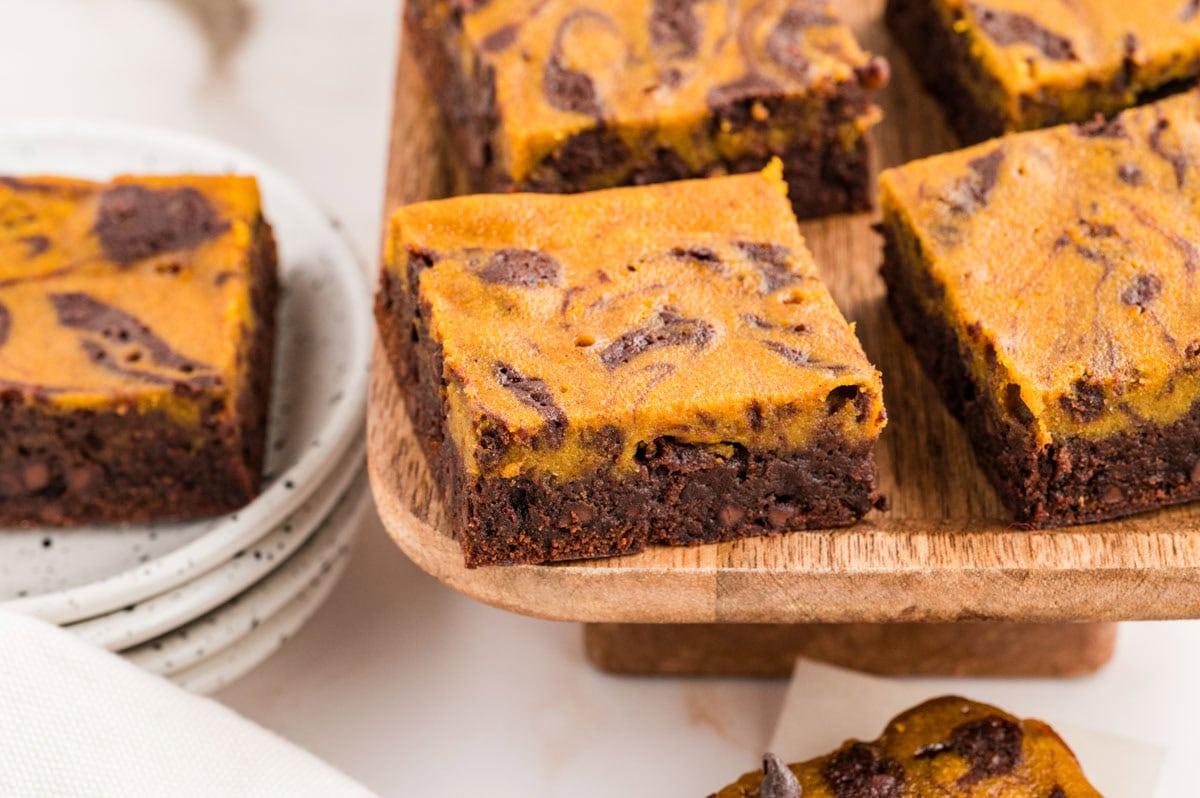 Pumpkin brownies on a wooden board with one on a stack of plates next to them.