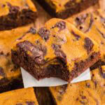 Pumpkin brownie with a bite taken out of it stacked on other pumpkin brownies on a wooden board.