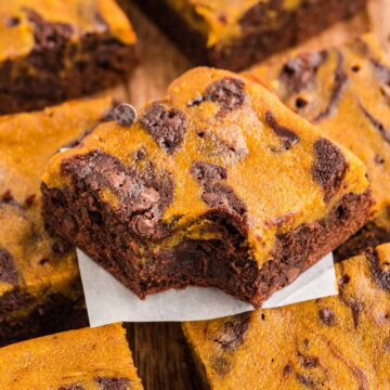 Pumpkin brownie with a bite taken out of it stacked on other pumpkin brownies on a wooden board.