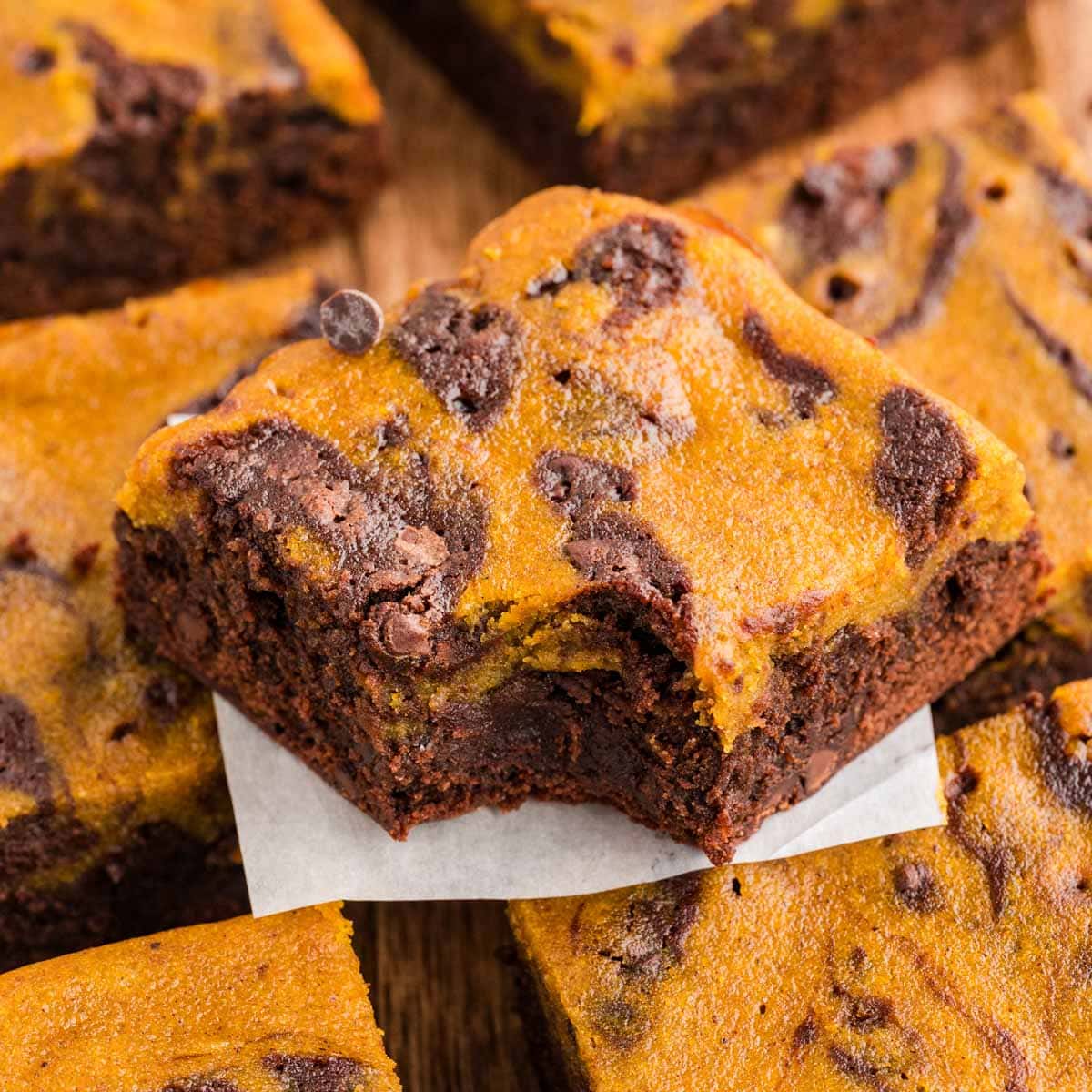 Pumpkin brownie with a bite taken out of it stacked on other pumpkin brownies on a wooden board.
