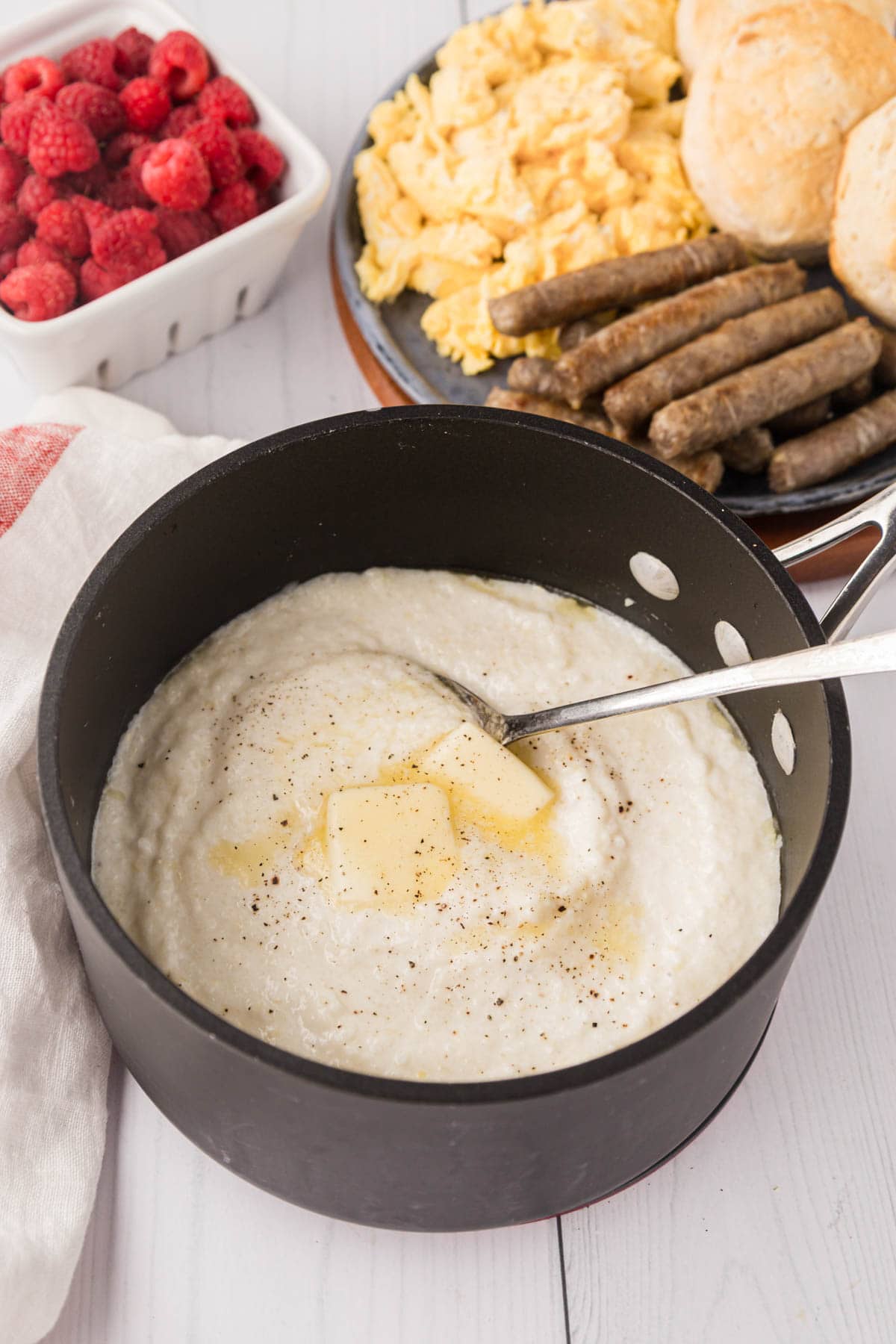 Grits in a pot with a platter of eggs, sausage and biscuits in the background.
