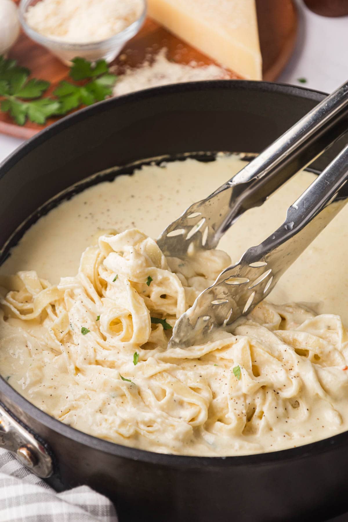 A black skillet filled with Alfredo sauce, using tongs to add in fettuccine noodles to toss and coat with sauce.