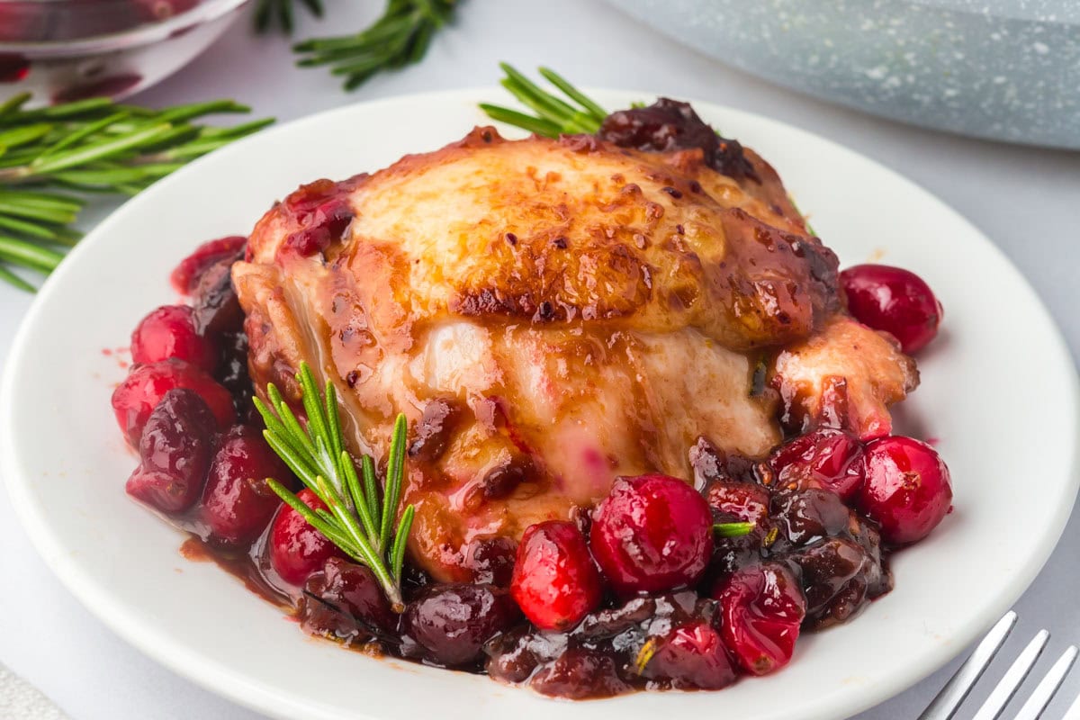 Balsamic cranberry chicken on a plate with cooked cranberries and a rosemary garnish.