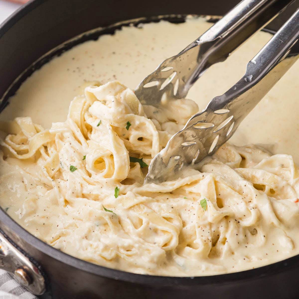 Homemade Alfredo sauce in a black skillet being tossed with fettuccine noodles.