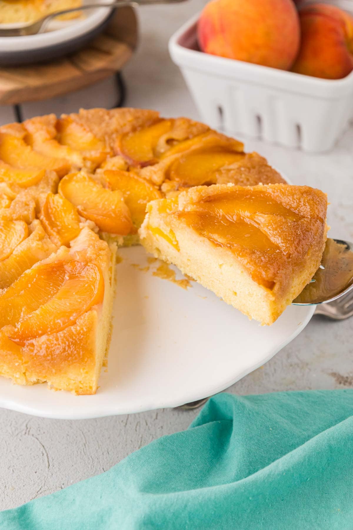 A cake stand with a peach upside down cake that has been cut and a slice being removed with cake server.