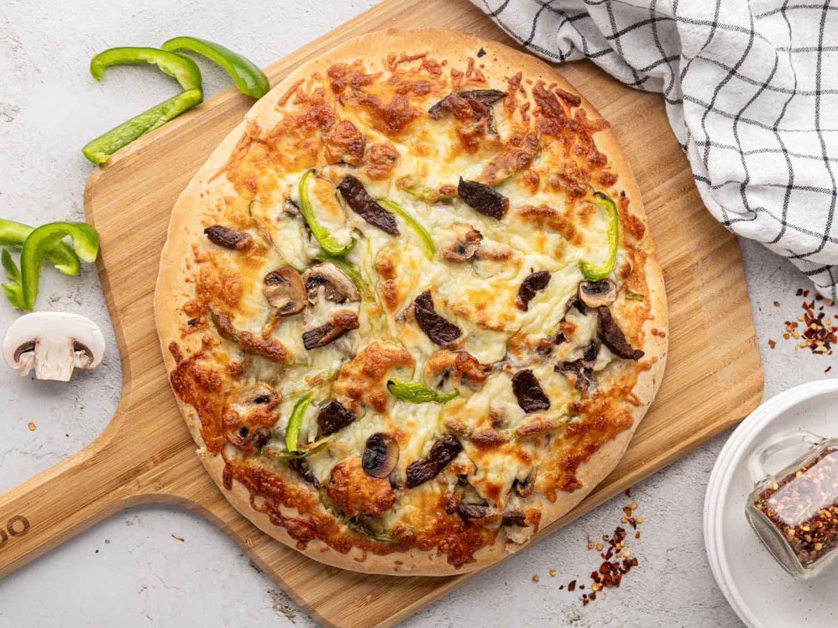 A Philly cheesesteak pizza with an Alfredo sauce base on a wooden pizza peel.
