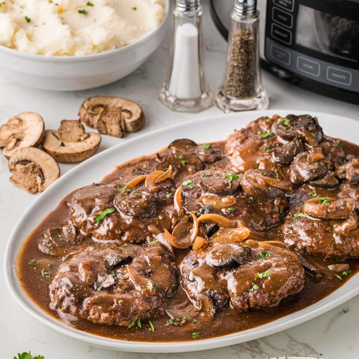 A white platter filled with Salisbury steak with a crockpot, mashed potatoes and salt and pepper in the background.