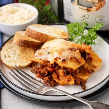 A serving ravioli lasagna made in the slow cooker on a white plate with garlic bread.