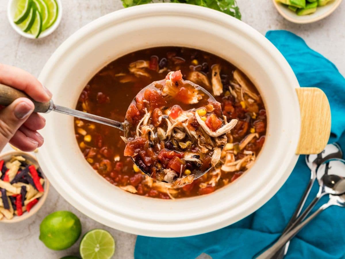 Overhead shot of slow cooker chicken tortilla soup with a hand holding a ladle of soup.