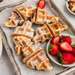 Croissant waffles served on a sheet pan with fresh strawberries and powdered sugar.