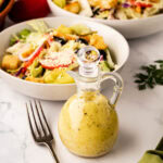 A salad dressing bottle filled with Olive Garden copycat dressing with a bowl and plated salad in the background.
