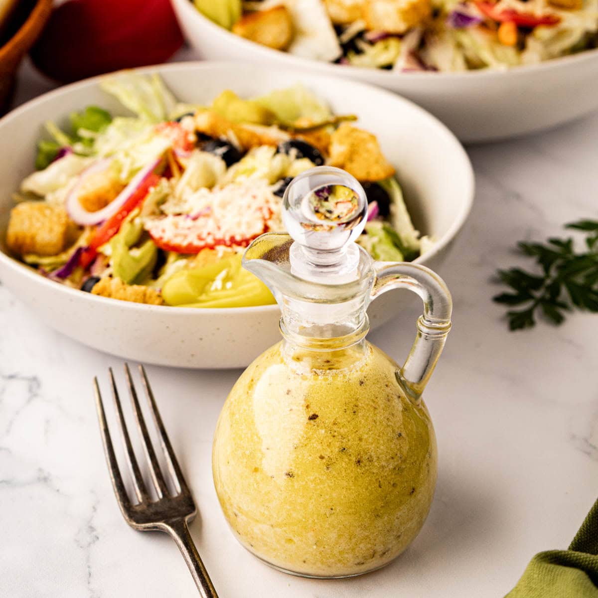 A salad dressing bottle filled with Olive Garden copycat dressing with a bowl and plated salad in the background.