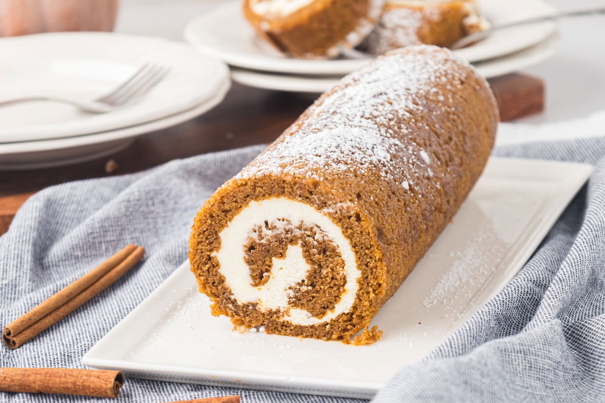 A whole pumpkin roll log cake on a white platter dusted with powdered sugar.