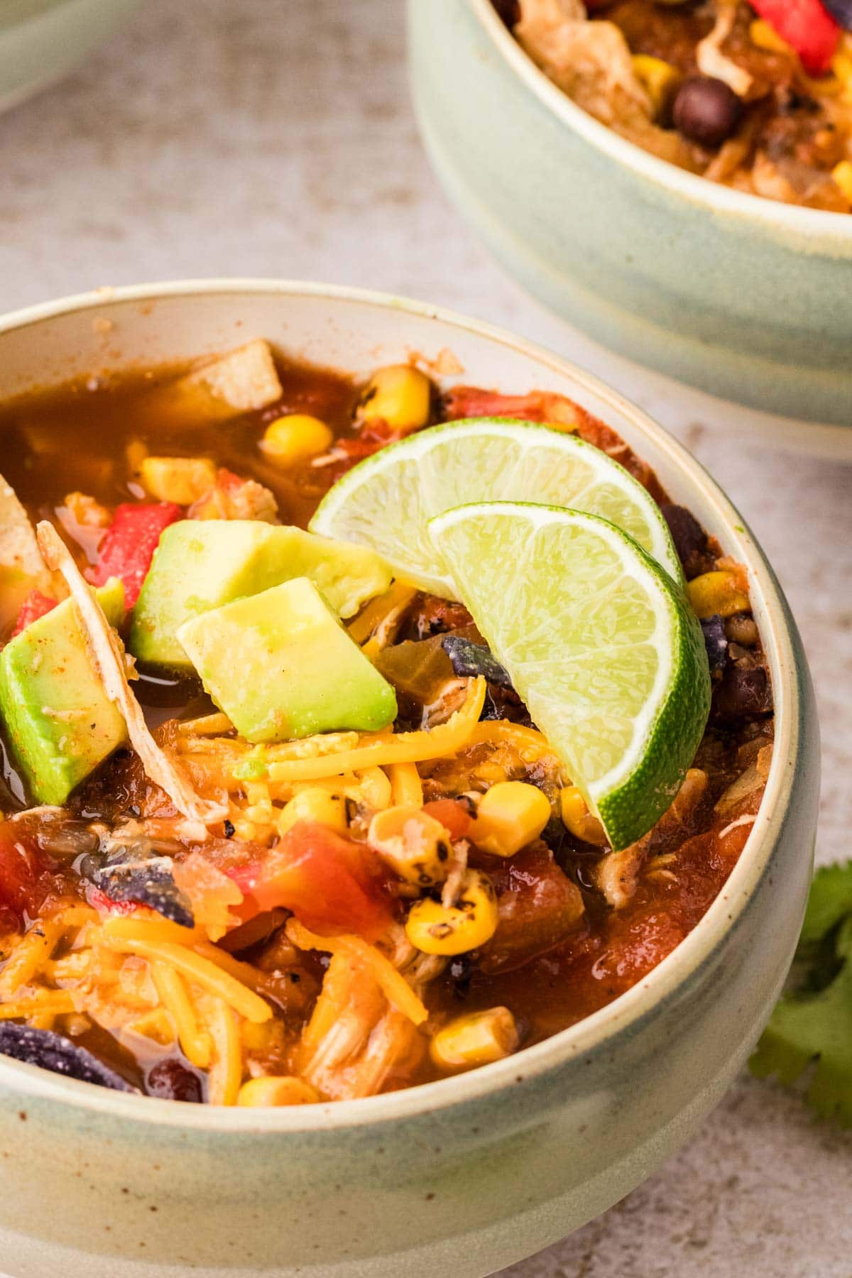 Close up of a bowl of chicken tortilla soup served with lime wedges and avocado on the top.