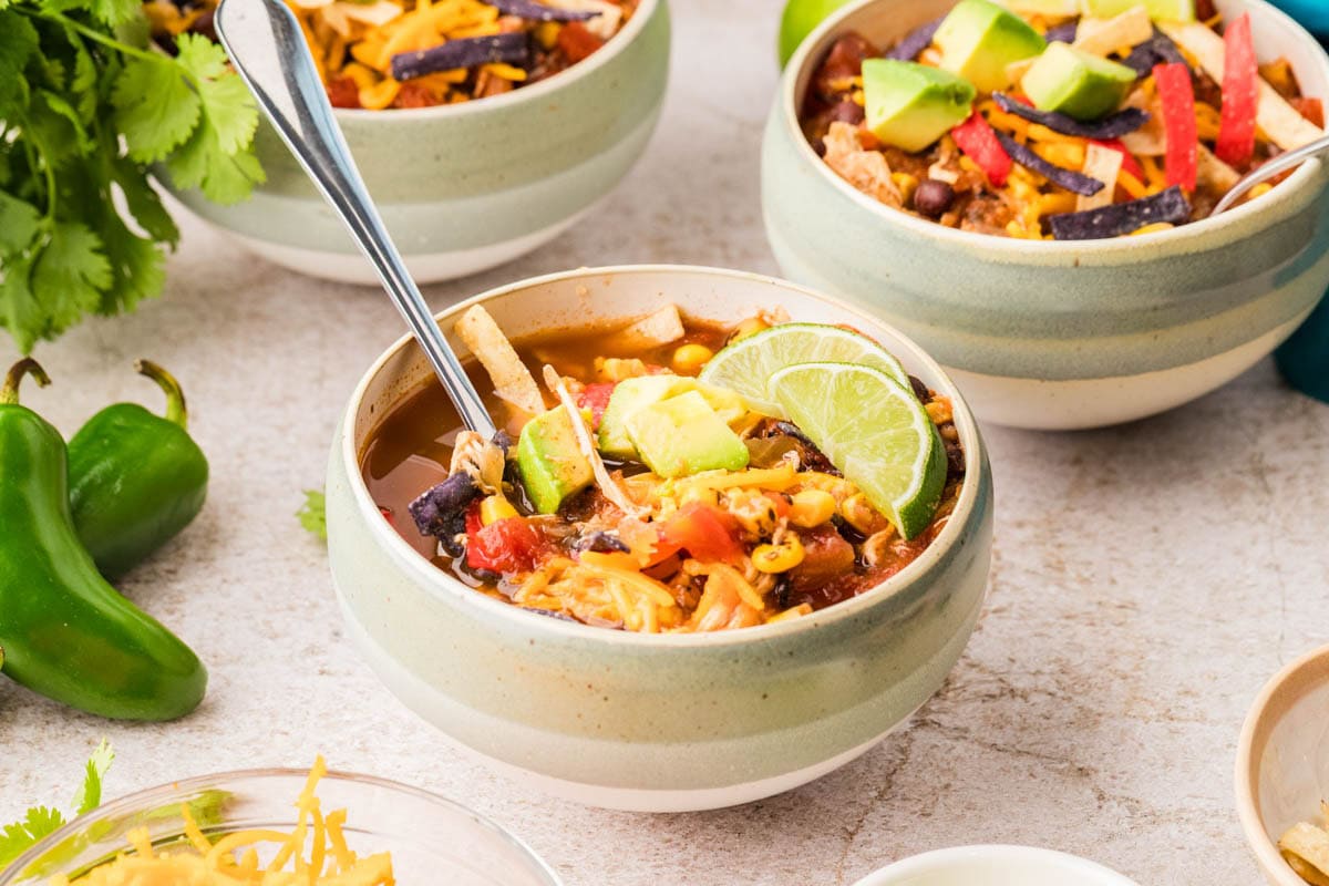 Bowl of slow cooker chicken tortilla soup with cheese, tortilla strips, avocado and lime wedges on top.