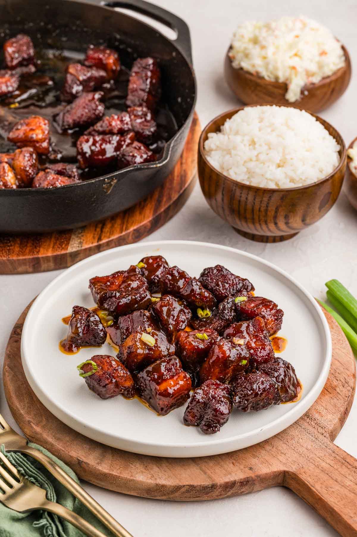 Smoked pork belly burnt ends served on a plate with some bowls of rice in the background.