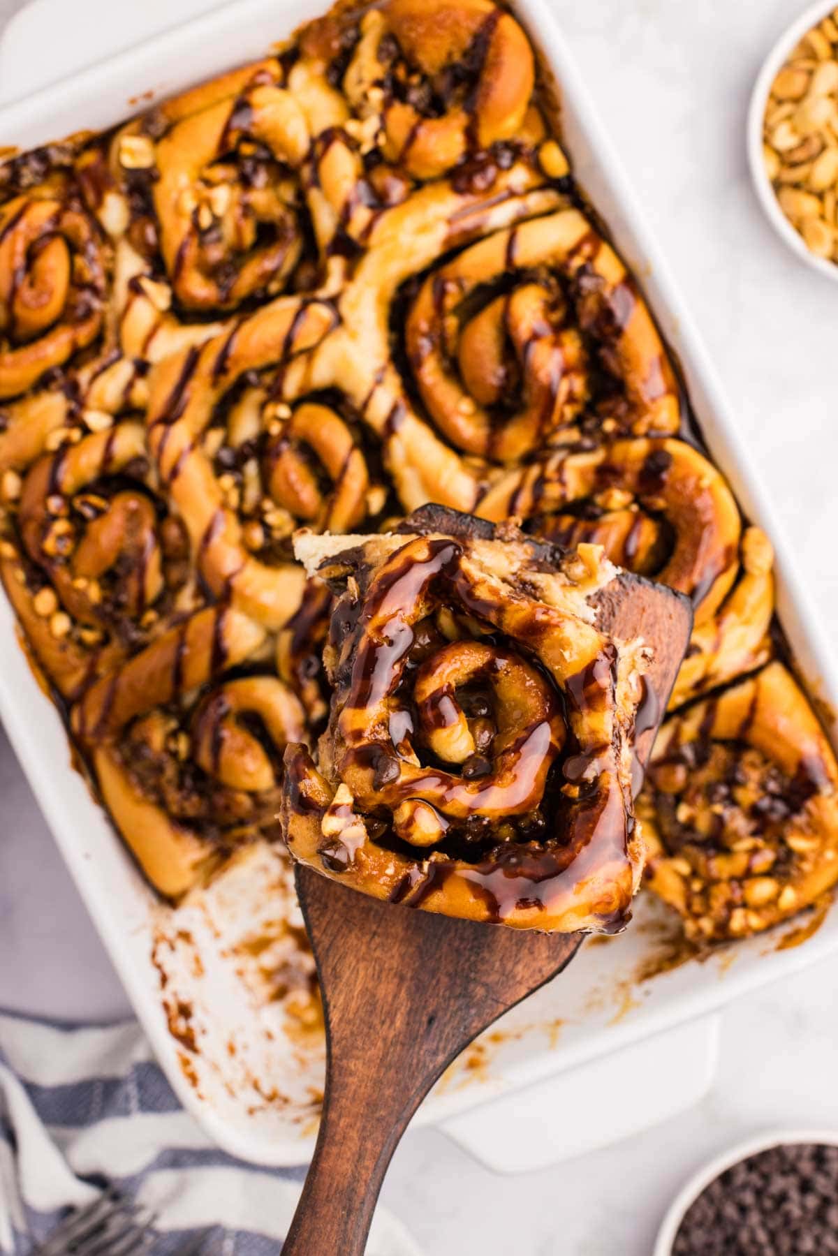 Wooden spatula with a snickers roll on it with more snickers rolls in the baking dish below.