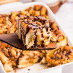 Wooden spatula with a homemade snickers roll on it with more rolls in a baking dish in the background.
