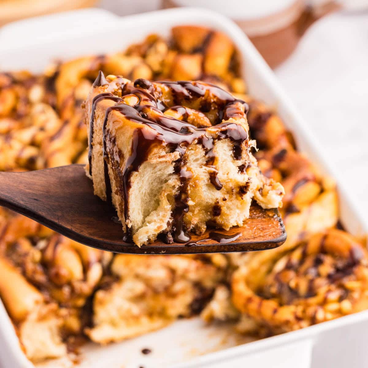 Wooden spatula with a homemade snickers roll on it with more rolls in a baking dish in the background.
