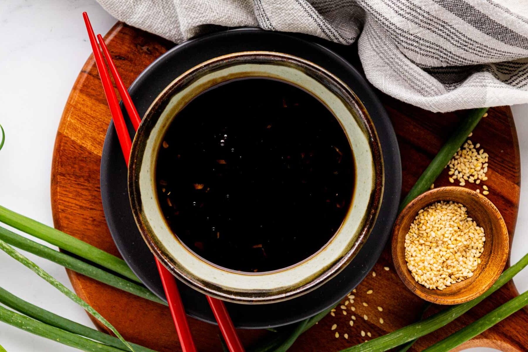 Teriyaki sauce in a bowl set on a wooden board with red chopstick, green onions and sesame seeds.