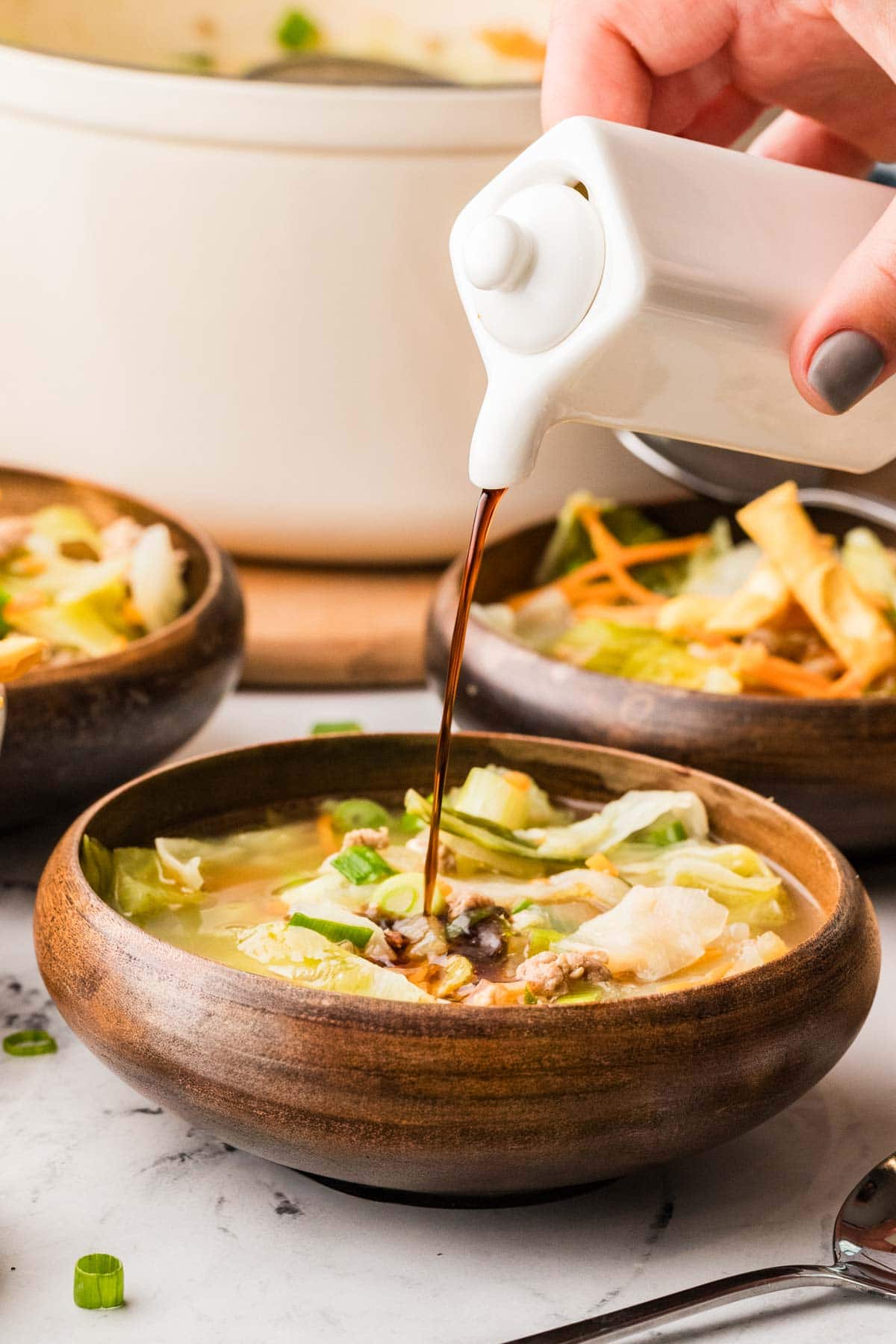 Hand pouring soy sauce into a wooden bowl filled with egg roll soup.