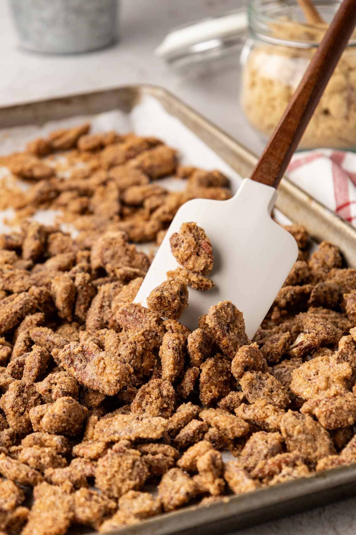 Pray Lee pecans on a baking sheet and a wooden spatula with a silicone tip, stirring the pecans.