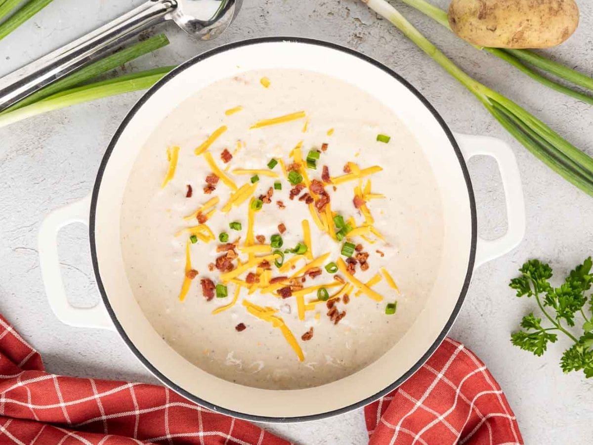 Creamy potato soup in a white Dutch oven topped with shredded cheese, bacon, and chives.
